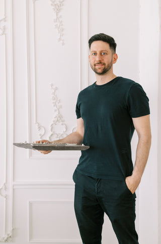 A man wearing a dark green crewneck t-shirt and matching dark pants stands indoors against a white paneled wall, holding a silver serving tray.