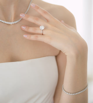 A close-up lifestyle shot of a woman's hand wearing a silver-toned ring with a round clear gemstone, along with matching diamond tennis necklaces and a bracelet.